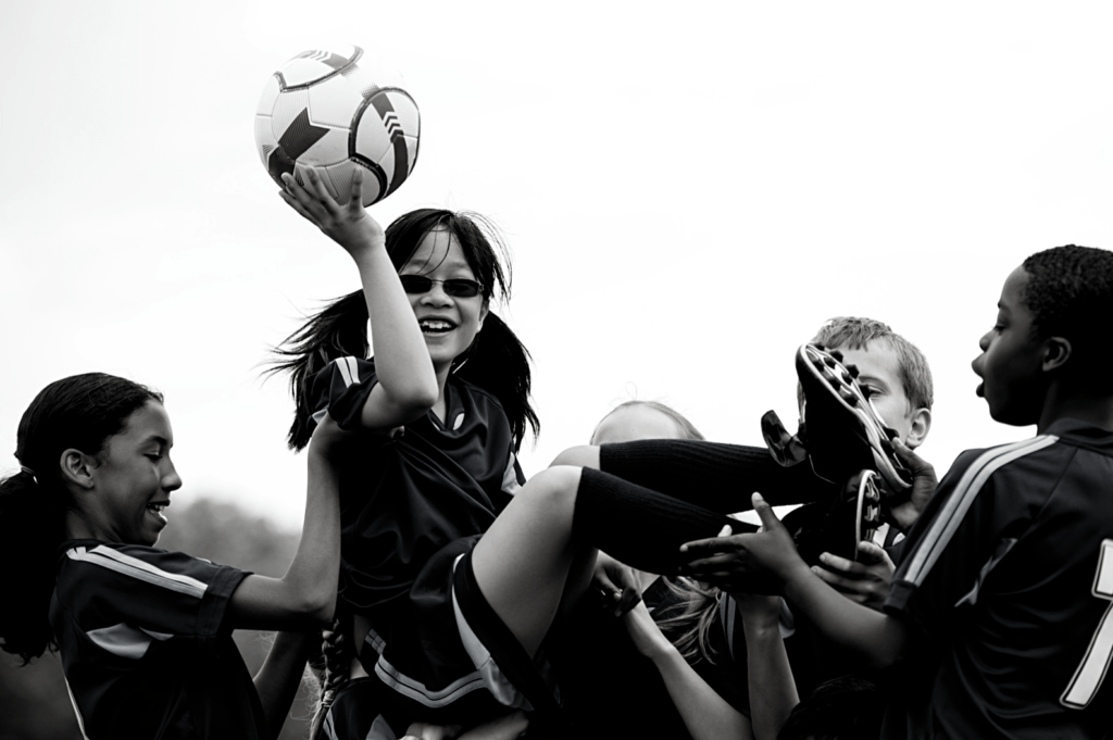 Varios niños y niñas de diferentes culturas festejan la celebración de un gol alzando a una niña por el aire que está sujetando un balón.